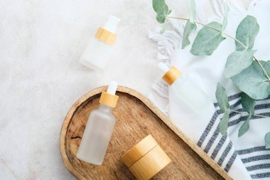 Doğal SPA kozmetik ürünleri, şeffaf cam ambalajlar, okaliptüs yaprakları ve banyodaki taş masanın üzerinde havlu. Düz yatış, üst görünüm.