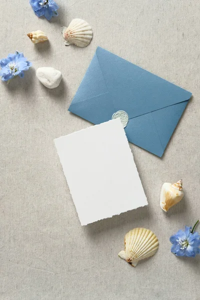 Nautical wedding invitation card and envelope with seashells and blue flowers on beige table. Wedding stationery set. Sea, marine, water style. Flat lay, top view.