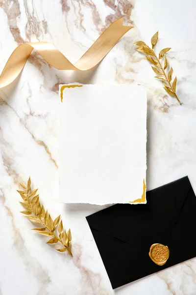 Blank paper card wedding invitation mockup, black envelope with wax stamp, silk ribbon, golden floral branches on gold marble table. Flat lay, top view.