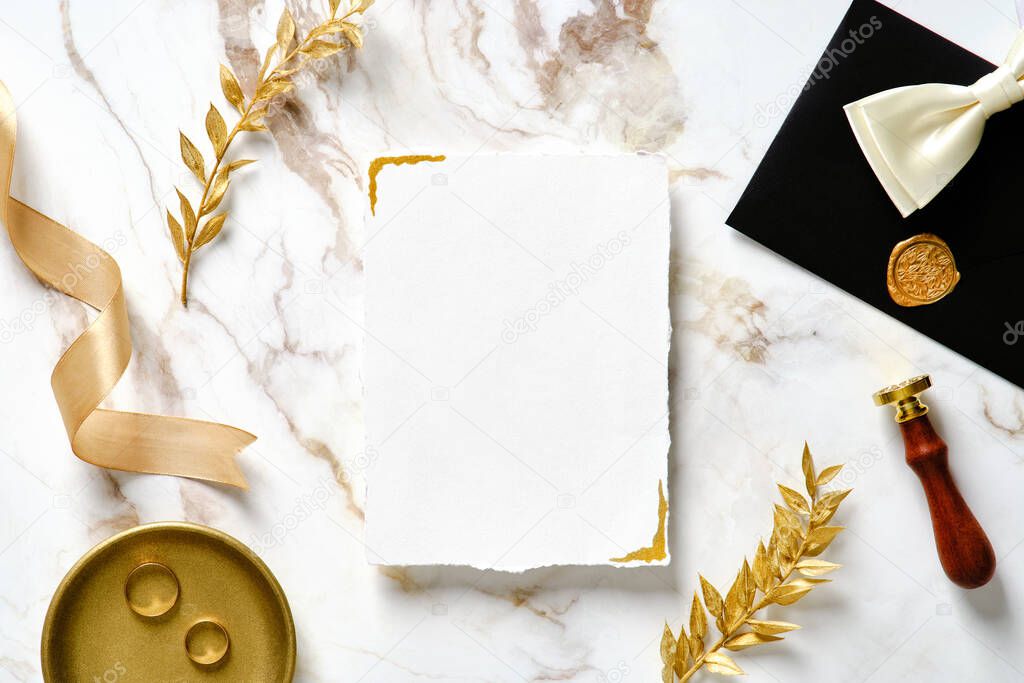 Wedding invitation card template. Top view blank paper card, golden rings, silk ribbon, wax seal stamp, golden branches on marble background. Luxury style. Flat lay.