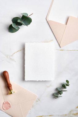 Boş kağıt kart düğün davetiyesi maketi, pastel pembe zarflar, balmumu mühür damgası, mermer masa üzerindeki okaliptüs yaprakları..