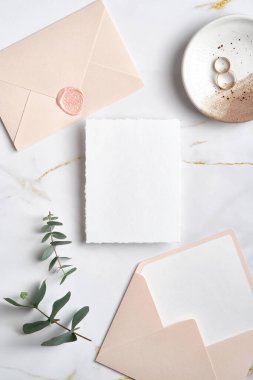 Düğün davetiyesi maketi, pastel pembe zarflar, altın yüzükler, mermer masadaki okaliptüs yaprakları. Zarif düğün kırtasiyesi seti. Düz yatış, üst görünüm.