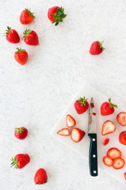 Çilekler, bütün ve kesilmiş, beyaz mermer kesme tahtasında bıçakla ve beyaz mermer üzerinde bir sürü fotokopi alanı ile. Üst Manzara. Dikey Sürüm