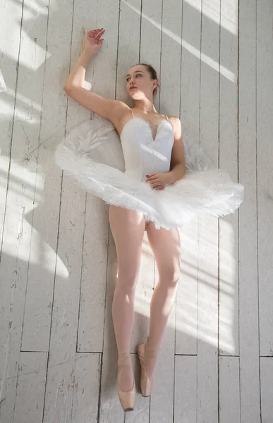 白い服とスカートのバレリーナは白い木製の床の上に延びた腕と長い足で横たわっています ストックフォト