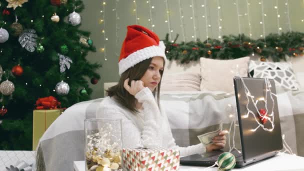 Kvinna i Santa hatt med dollarsedel i händerna tänka och skriva på laptop, val av online shopping eller betalning vid jul — Stockvideo