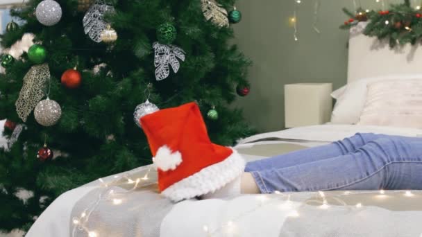 Gambe femminili con cappello di Babbo Natale sul letto con luci natalizie festive ballano un po 'a casa, umore natalizio senza volto — Video Stock