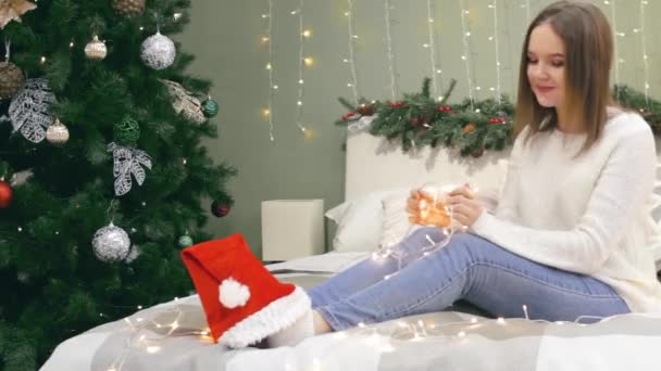 Mulher com chapéu de Papai Noel nas pernas e luzes de xmas nas mãos dançam um pouco na cama no quarto com árvore de natal em casa — Vídeo de Stock
