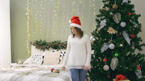 Joven mujer bonita en el sombrero de santa decorar el árbol de Navidad por la decoración de la bola de Navidad en la habitación de la cama con regalos en casa — Vídeos de Stock