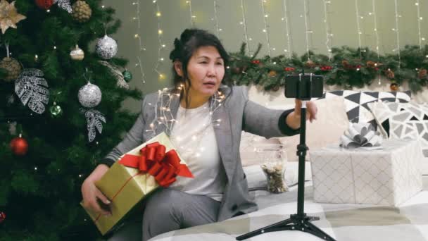 クリスマスライトと金のギフトボックスを持つシニアアジアの女性は 自宅で寝室でのビデオ通話 幸せなお祝いだけで クリスマスや新年のお祝いオンライン会議でセルフィーや電話で話す — ストック動画