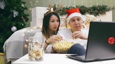 Noel 'de dizüstü bilgisayarında patlamış mısır filmi olan çift Noel Baba şapkalı beyaz adam ve geyik boynuzlu Asyalı kadın.