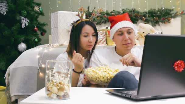 Para z popcornem oglądaj film na laptopie na Boże Narodzenie, biały mężczyzna w czapce Świętego Mikołaja i azjatka kobieta z rogami reniferów — Wideo stockowe
