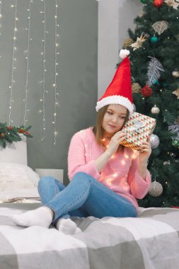 Noel Baba şapkalı kadın elinde hediye kutusuyla yatakta oturur. Yatak odasının iç dekorasyonu Noel ağacı ve Noel ışıkları ile süslenir.