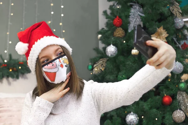 Mulher em santa chapéu e máscara de tecido com design xmas leva selfie ou vídeo chamada por telefone em casa, novo normal de Natal — Fotografia de Stock