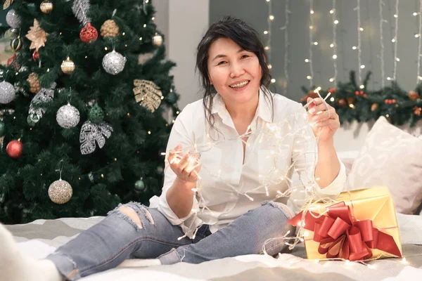 Senior woman on bed with xmas lights, attractive elderly smiling lady with garland in hands at home at christmas holiday