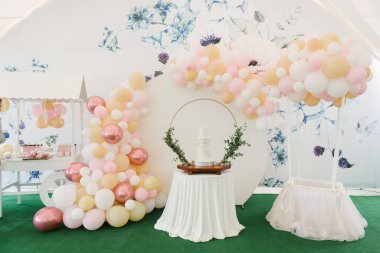 Balonlu ve çikolatalı güzel bir fotoğraf bölgesinin arka planında yeşil dallar olan bir çocuğun vaftiz töreni için büyük, üç katlı beyaz bir pasta.