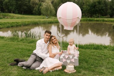 Güzel küçük bir kız, anne ve babasıyla birlikte bir sepetin içinde balonla ve bir pastayla ilk doğum gününü kutluyor.