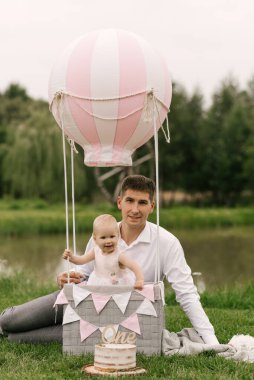 Güzel küçük bir kız babasının yanında bir sepetin içinde balonla ve bir pastayla ilk doğum gününü kutluyor.