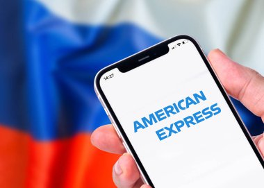Antalya, Türkiye - 06 Mart 2022: Amerikan Ekspres logosu ile akıllı telefon tutan adam bulanık Rus bayrağının önünde duruyor