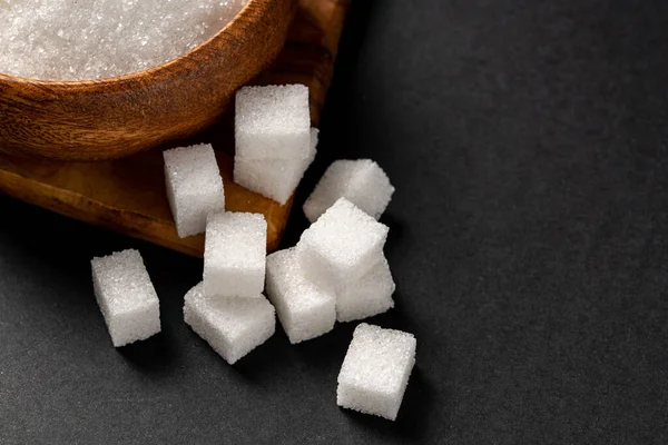 Granulated sugar and sugar cubes in wooden bowl on dark background