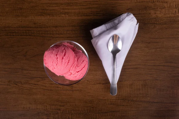 Strawberry flavored ice cream served in a glass bowl next to a spoon on a white cloth. Top photo.