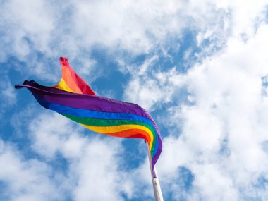 Rüzgarda homo bir gurur bayrağı dalgalanıyor. Arkasında kabarık bulutlar olan mavi bir gökyüzü..