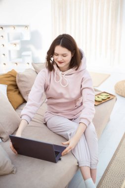 Genç gülümseyen bir kadın kanepede oturuyor ve dizüstü bilgisayarına bakıyor.