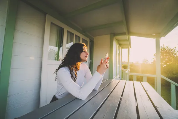 日落时坐在平房阳台上拿着手机的女人 — 图库照片