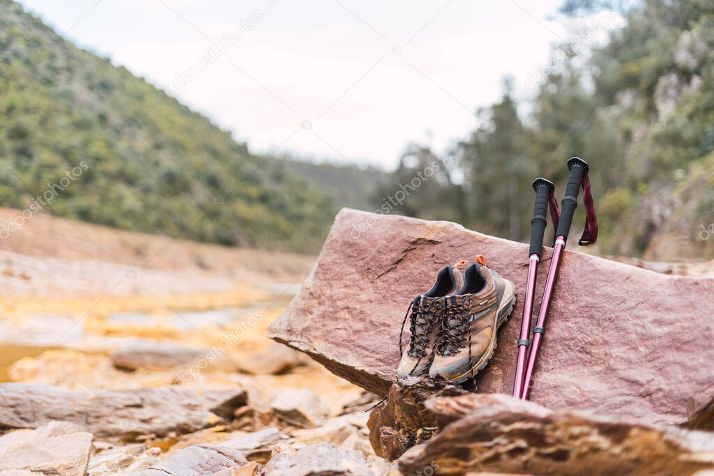 Close-up of hiking boots and trekking poles on a stone with copy space, in the background green mountains and the flowing water of the Rio Tinto.