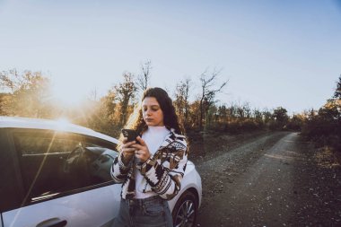 Ağaçlar arasındaki yolda bir arabanın yanındaki telefona bakan kadın.