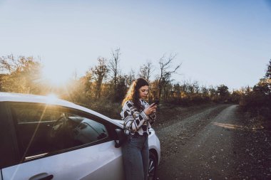 Ağaçlar arasındaki yolda bir arabanın yanındaki telefona bakan kadın.