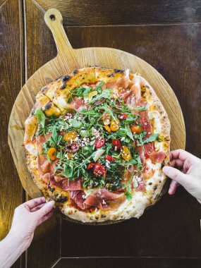 İki elle jambonlu, roka ve parmesanlı pizzayı ahşap bir tabaktan al.