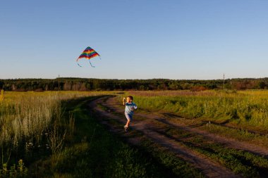 Bir çocuk elinde gökkuşağı uçurtmasıyla tarlaya doğru koşuyor.