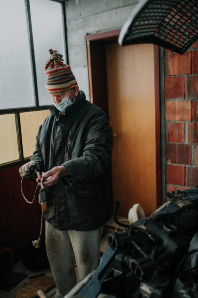 Старик в защитной маске в гараже ремонтирует свою старую машину.