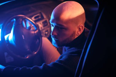 Mavi ışıkla aydınlatılmış araba kullanan kel bir gencin portresi.