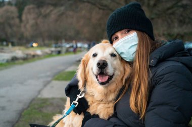 Parktaki bankta köpeğini kucaklayan kadın, siyah bir maske takıyor.