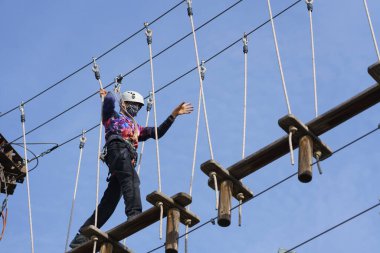 Çocuk, güvenli ip ve kaskla bir macera parkında tahta köprüde yürürken eğleniyor.