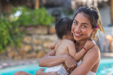 Güzel bir Latin annenin portresi havuz başında bebeğine sarılıyor.