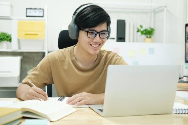 Genç erkek öğrenci evde ders çalışıyor. Bilgisayarı kullanıyor ve internetten öğreniyor.