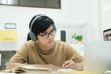 Genç erkek öğrenci evde ders çalışıyor. Bilgisayarı kullanıyor ve internetten öğreniyor.