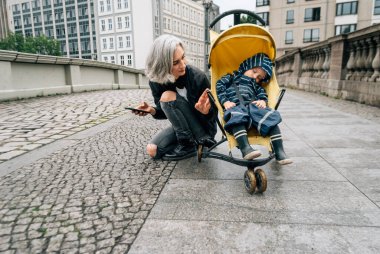 Anne, küçük oğlu kaldırımda otururken bebek arabasına bakıyor.