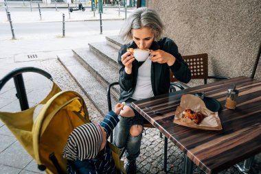 Kadın kahve içer ve kafede çörek yer. Bebek arabasının yanında çocuk var.  