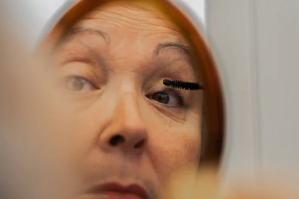 older woman painting her eyelashes in front of a mirror