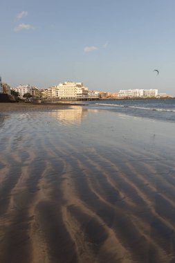 Gün batımında deniz manzarası, okyanus kıyısında ıslak sarı kumsal ve arka planda resimli bir balıkçı köyü. Playa Grande, El Medano, Tenerife, Kanarya Adaları. İspanya