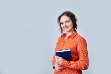 Elinde not defteriyle gülümseyen orta yaşlı bir iş kadını. Mesajlaşmak için yeri olan gri bir duvara yaslanmış..