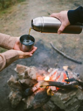 a man pours tea from a thermos near a fire