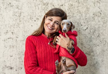 Latin kadın köpeğini tutuyor, ikisi de kırmızı giyinmiş ve kameraya bakıyor.