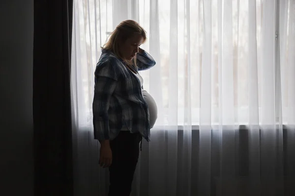 Pregnant woman in home clothes stands at the window