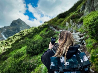 Sırt çantalı genç bir turist dağlarda telefonla fotoğraf çekiyor.