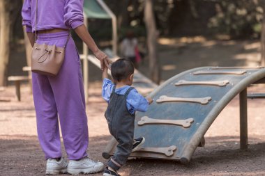 Küçük çocuk parkta oynuyor. Annesinin elini tutarak rampadan tırmanıyor.