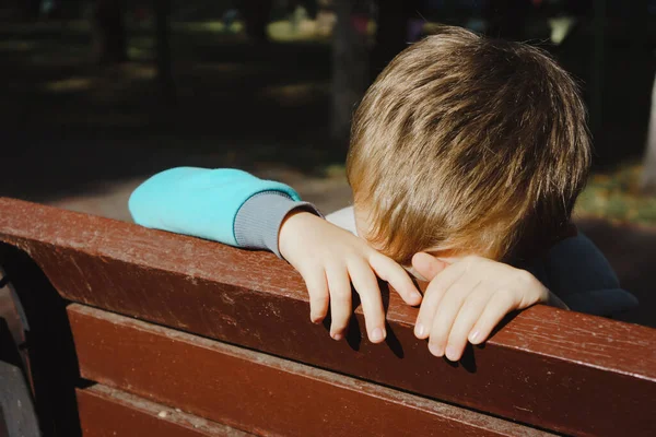 Sad boy hides his face behind a park bench outdoors close-up